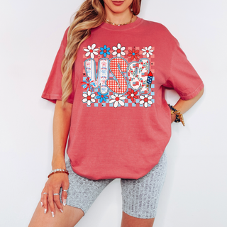 Woman wearing a faded red oversized t-shirt with bold “USA” lettering in gingham and floral prints, surrounded by red, white, and blue daisies, styled with light gray biker shorts.