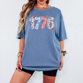 Woman wearing a beige oversized graphic t-shirt that reads “1776” in red gingham and floral prints, styled with casual biker shorts and summer accessories.