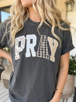 A woman wearing a dark gray t-shirt with the word 'PRAY' printed across the chest in a patchwork pattern of floral, striped, and plaid lettering.