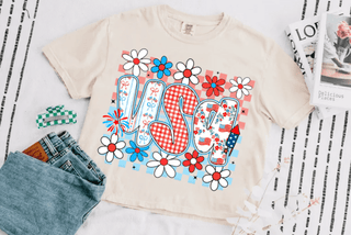 Woman wearing a faded red oversized t-shirt with bold “USA” lettering in gingham and floral prints, surrounded by red, white, and blue daisies, styled with light gray biker shorts.