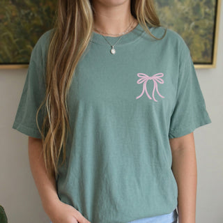 Person wearing a green t-shirt with a pink bow design and denim shorts indoors.