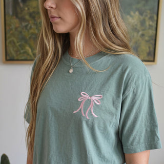 Woman wearing a green t-shirt with a pink bow design in a casual setting.