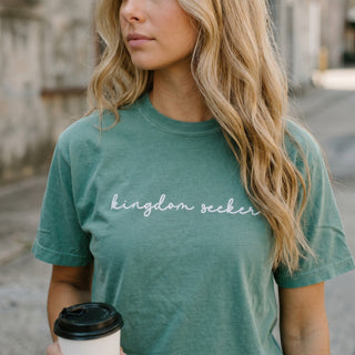 Woman wearing a teal t-shirt with 'kingdom seeker' text, holding a coffee cup outdoors.