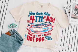 Woman wearing a cream oversized t-shirt with a red, white, and blue hot dog graphic and patriotic text that says "You Look Like the 4th of July – Makes Me Want a Hot Dog Real Bad," styled with black biker shorts.