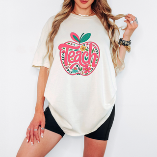 Woman wearing an oversized cream graphic tee with a retro floral apple design and the word ‘Teach’, styled with black biker shorts